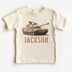 May include: A beige short-sleeved t-shirt with a camouflage tank printed on the front. The text "JACKSON" is printed below the tank in brown.
