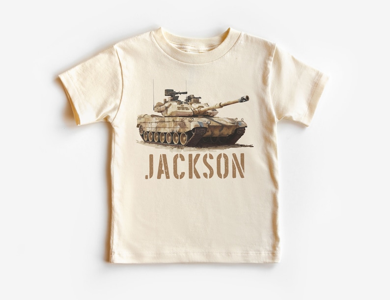 May include: A beige short-sleeved t-shirt with a camouflage tank printed on the front. The text "JACKSON" is printed below the tank in brown.