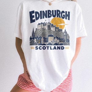 Pu&ograve; includere: Una t-shirt bianca con una stampa grafica dello skyline di Edimburgo, in Scozia. Il design include il testo "EDINBURGH" in blu scuro, un sole giallo e il testo "SCOTLAND" con due linee orizzontali gialle su entrambi i lati.