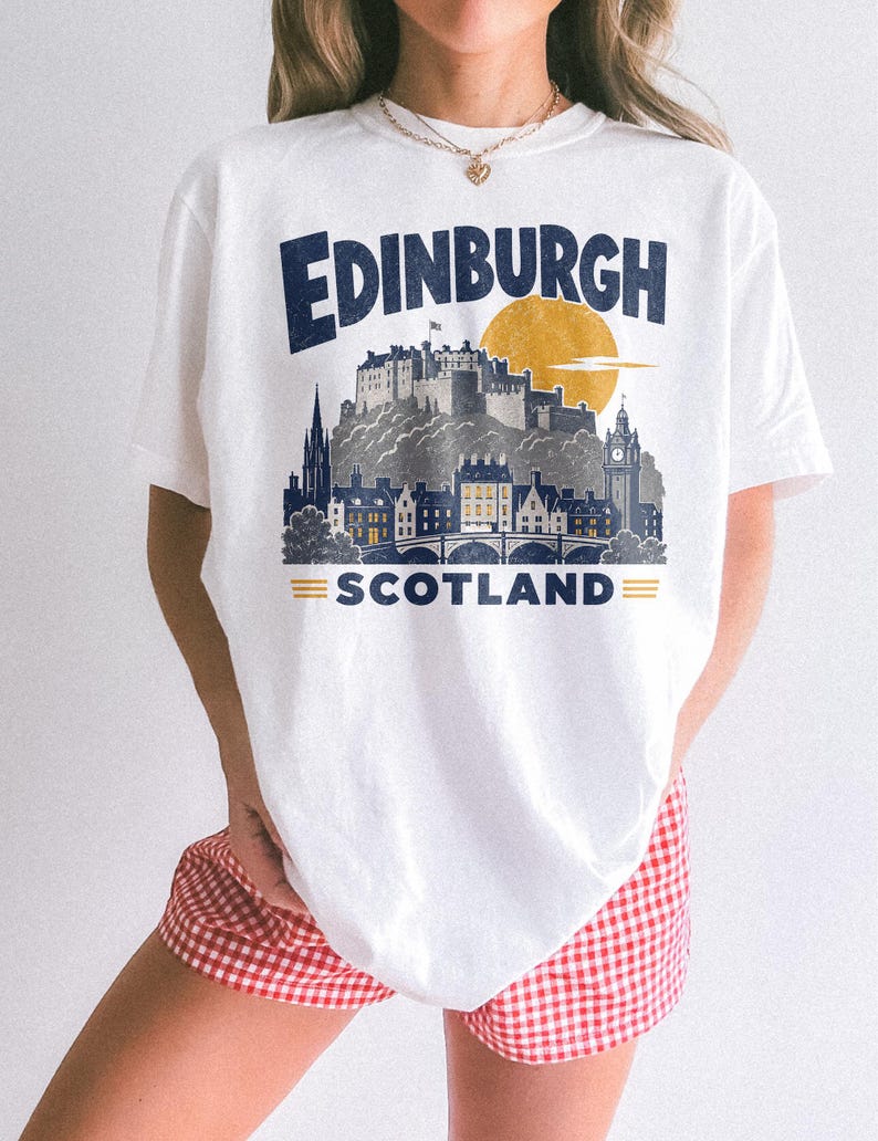 Pu&ograve; includere: Una t-shirt bianca con una stampa grafica dello skyline di Edimburgo, in Scozia. Il design include il testo "EDINBURGH" in blu scuro, un sole giallo e il testo "SCOTLAND" con due linee orizzontali gialle su entrambi i lati.