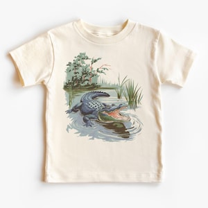 Op de afbeelding: Een crèmekleurig T-shirt met een afbeelding van een alligator in een moeras. De alligator is blauw en groen en heeft zijn bek open.