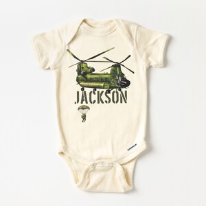 May include: A cream-colored baby onesie with a green helicopter and a parachutist printed on the front. The text "JACKSON" is printed below the helicopter.