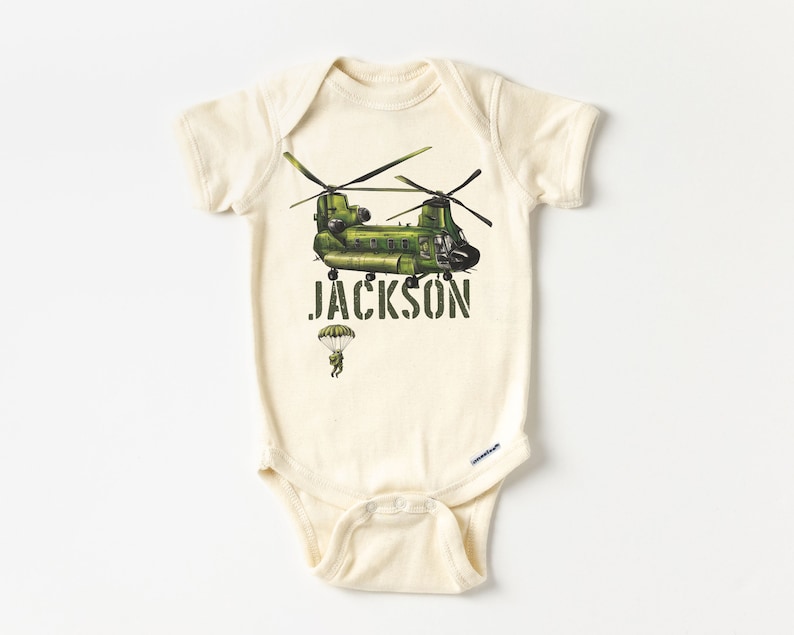 May include: A cream-colored baby onesie with a green helicopter and a parachutist printed on the front. The text "JACKSON" is printed below the helicopter.