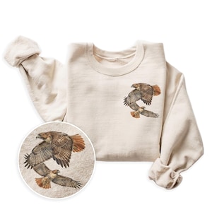 May include: Cream-colored sweatshirt featuring a detailed illustration of three hawks in flight. The artwork showcases the birds with brown and orange feathers. The design is repeated in a circular inset, highlighting the intricate details of the artwork.