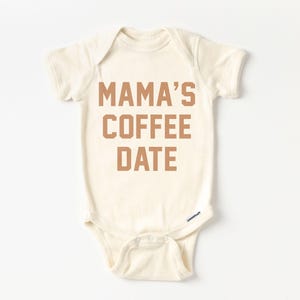 Puede incluir: Un body de beb&eacute; de manga corta en color crema con el texto MAMA'S COFFEE DATE impreso en una fuente marr&oacute;n de estilo bloque en el pecho. La prenda cuenta con un cuello redondo y un cierre de botones a presi&oacute;n en la parte inferior.