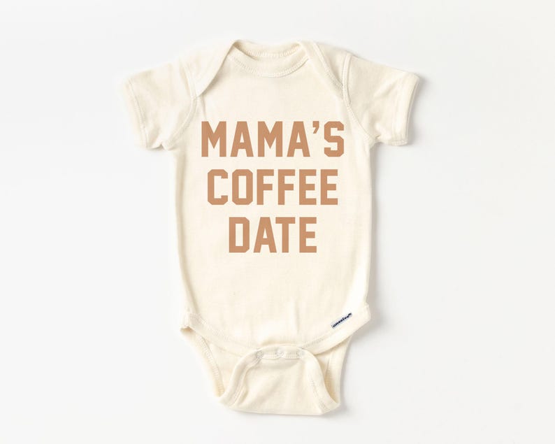 Puede incluir: Un body de beb&eacute; de manga corta en color crema con el texto MAMA'S COFFEE DATE impreso en una fuente marr&oacute;n de estilo bloque en el pecho. La prenda cuenta con un cuello redondo y un cierre de botones a presi&oacute;n en la parte inferior.