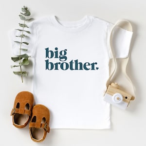 May include: White t-shirt with the text "big brother." in dark blue letters. The shirt is laying flat on a white surface with a pair of brown leather shoes and a small wooden toy camera.