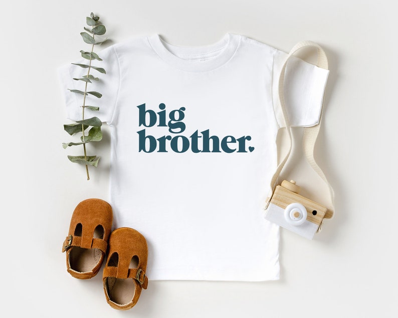 May include: White t-shirt with the text "big brother." in dark blue letters. The shirt is laying flat on a white surface with a pair of brown leather shoes and a small wooden toy camera.