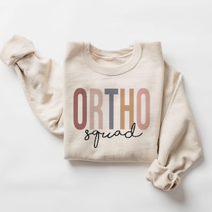 May include: A beige sweatshirt with the text "ORTHO squad" in colourful letters. The letters are in shades of pink, brown, beige, and blue.