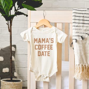 Puede incluir: Un body de beb&eacute; de manga corta en color crema con el texto MAMA'S COFFEE DATE impreso en una fuente marr&oacute;n negrita sin remates en el pecho. La prenda incluye un cuello redondo y cierres de presi&oacute;n met&aacute;licos en la entrepierna.