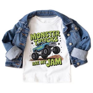 Puede incluir: Camiseta blanca con un gr&aacute;fico de cami&oacute;n monstruo azul y el texto "MONSTER TRUCKS ARE MY JAM" en verde y negro. Una chaqueta vaquera azul est&aacute; sobre la camiseta.