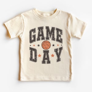 May include: A cream coloured t-shirt with a black and orange basketball graphic and the text "GAME DAY" in a vintage style font.