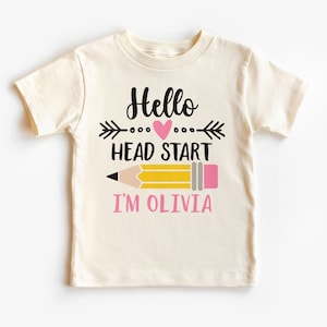 May include: Cream-colored t-shirt with the text "Hello Head Start I'm Olivia" in black and pink. The design includes a pink heart, an arrow, and a yellow pencil with a pink eraser. A cute shirt for a child.