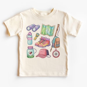 K&ouml;nnte beinhalten: Cremefarbenes T-Shirt mit einer farbenfrohen Illustration von Outdoor-Abenteuer-Ausr&uuml;stung. Das Design umfasst Fernglas, eine Karte, Wanderschuhe, einen Rucksack, einen Kompass, eine Wasserflasche, eine Taschenlampe, ein Glas mit einem Marienk&auml;fer und eine Baseballkappe.