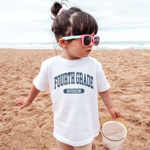 May include: A white t-shirt with the text "FOURTH GRADE HUDSON" in navy blue. The child is wearing pink sunglasses and holding a yellow bucket on a sandy beach.
