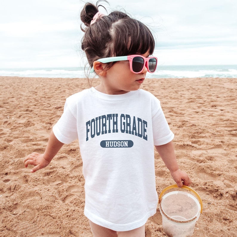 May include: A white t-shirt with the text "FOURTH GRADE HUDSON" in navy blue. The child is wearing pink sunglasses and holding a yellow bucket on a sandy beach.