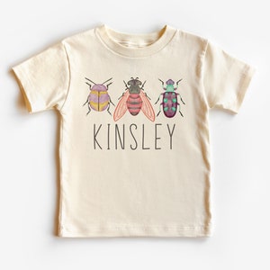 以下が含まれることがあります： クリーム色の半袖Tシャツ。水彩画の虫3匹と「Kinsley」という名前が前面にプリントされています。