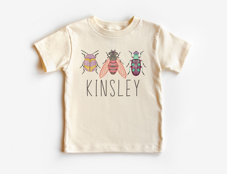 以下が含まれることがあります： クリーム色の半袖Tシャツ。水彩画の虫3匹と「Kinsley」という名前が前面にプリントされています。