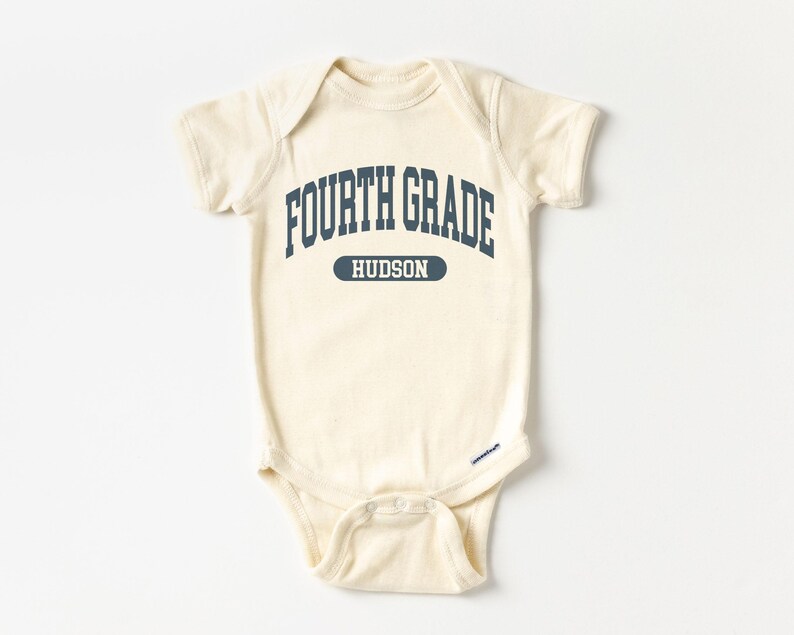 May include: Cream-colored baby bodysuit with the text "FOURTH GRADE" in navy blue arched lettering. Below the text is the name "HUDSON" in a navy blue rectangle. The bodysuit has short sleeves and a snap closure.
