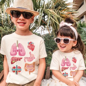 K&ouml;nnte beinhalten: Zwei Kinder tragen wei&szlig;e T-Shirts mit bunten Illustrationen von menschlichen Organen. Die Shirts zeigen die W&ouml;rter "Lungen", "Herz", "Magen", "Leber", "Nieren" und "D&auml;rme".