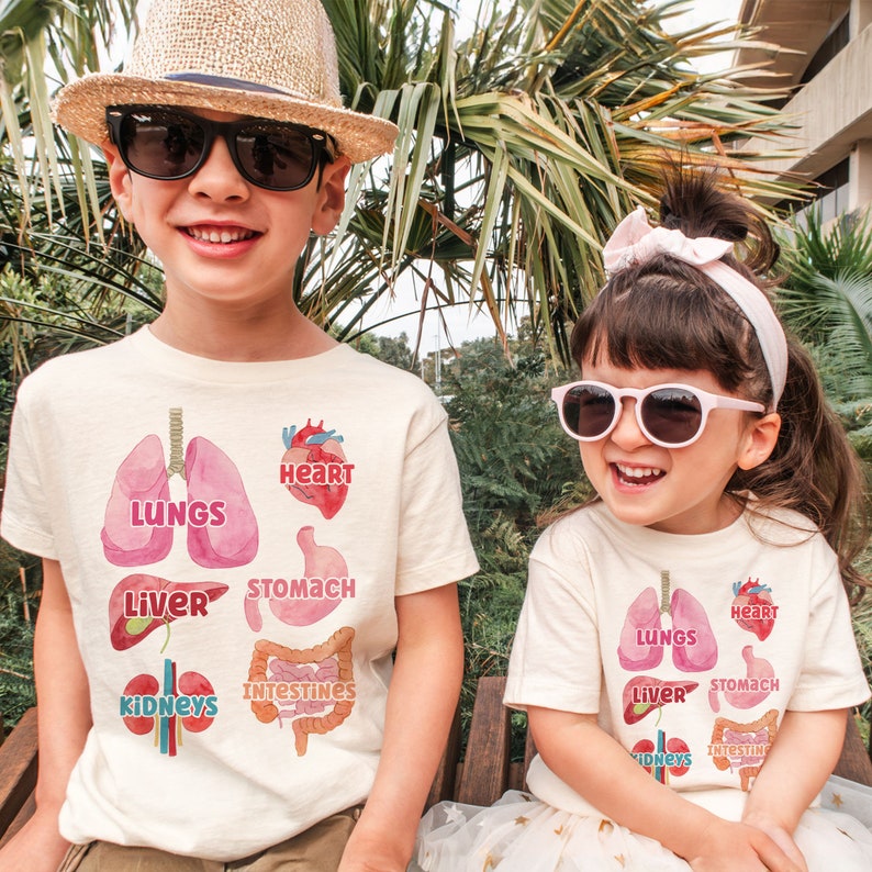 K&ouml;nnte beinhalten: Zwei Kinder tragen wei&szlig;e T-Shirts mit bunten Illustrationen von menschlichen Organen. Die Shirts zeigen die W&ouml;rter "Lungen", "Herz", "Magen", "Leber", "Nieren" und "D&auml;rme".