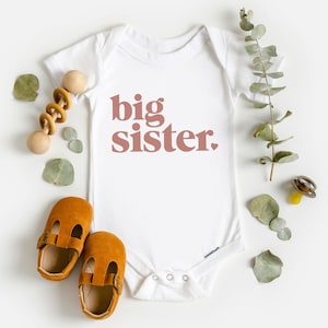 May include: White baby onesie with the words "big sister" printed in pink on the front. The onesie is surrounded by a wooden rattle, brown baby shoes, and eucalyptus leaves.