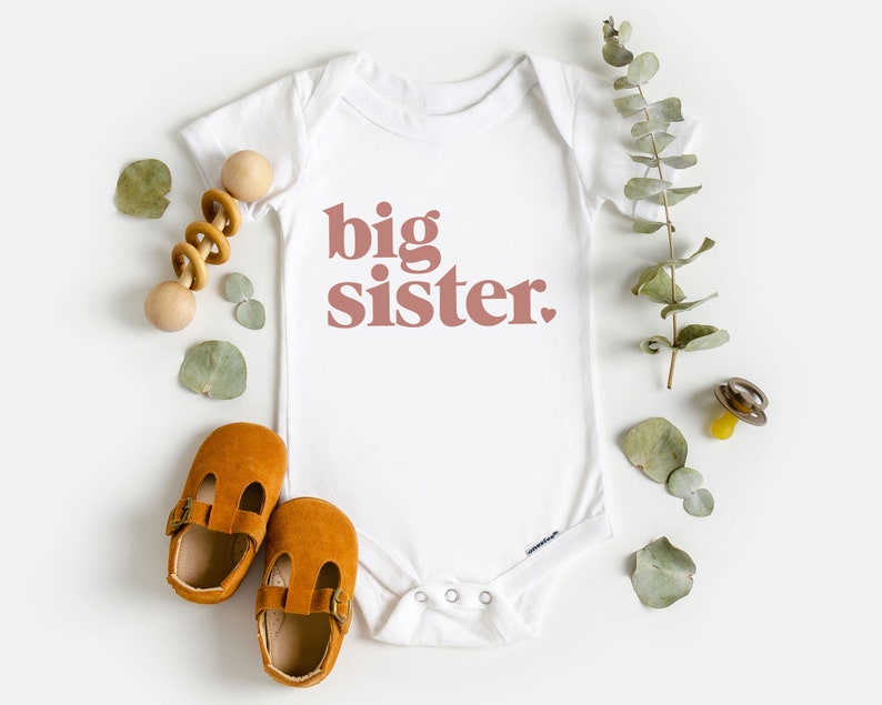 May include: White baby onesie with the words "big sister" printed in pink on the front. The onesie is surrounded by a wooden rattle, brown baby shoes, and eucalyptus leaves.