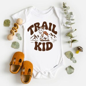 Peut inclure: Body b&eacute;b&eacute; blanc avec un motif marron &laquo;&nbsp;TRAIL KID&nbsp;&raquo; avec une ch&egrave;vre et des montagnes. L'image comprend &eacute;galement des chaussures de b&eacute;b&eacute; marron et un jouet en bois.