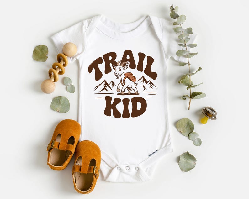 Peut inclure: Body b&eacute;b&eacute; blanc avec un motif marron &laquo;&nbsp;TRAIL KID&nbsp;&raquo; avec une ch&egrave;vre et des montagnes. L'image comprend &eacute;galement des chaussures de b&eacute;b&eacute; marron et un jouet en bois.