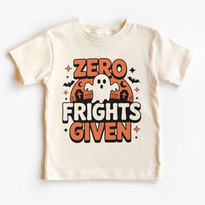 May include: Cream-colored t-shirt with the text "ZERO FRIGHTS GIVEN" in orange and white, featuring a ghost, bats, and a tombstone design. A fun Halloween-themed graphic tee for kids.
