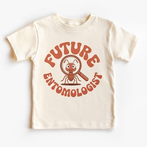 Puede incluir: Camiseta color crema con una hormiga de dibujos animados sosteniendo una lupa. El texto "FUTURE ENTOMOLOGIST" está impreso en un diseño circular alrededor de la hormiga en marrón rojizo.