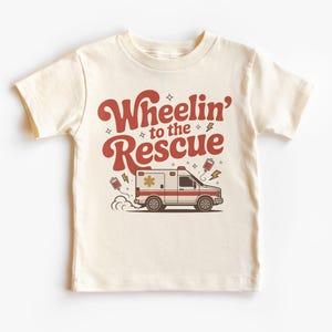 Puede incluir: Camiseta color crema para niños con la frase "Wheelin' to the Rescue" en rojo con tipografía retro. Debajo, una ambulancia de dibujos animados con detalles rojos y blancos, humo y rayos.
