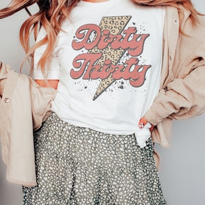 May include: White t-shirt with a red and yellow graphic that says "Dirty Thirty" with a leopard print lightning bolt.
