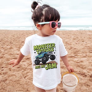 Puede incluir: Camiseta blanca con un gr&aacute;fico de un cami&oacute;n monstruo azul y el texto "MONSTER TRUCKS ARE MY JAM" en verde y negro. El ni&ntilde;o en la playa de arena lleva la camiseta y gafas de sol rosas.