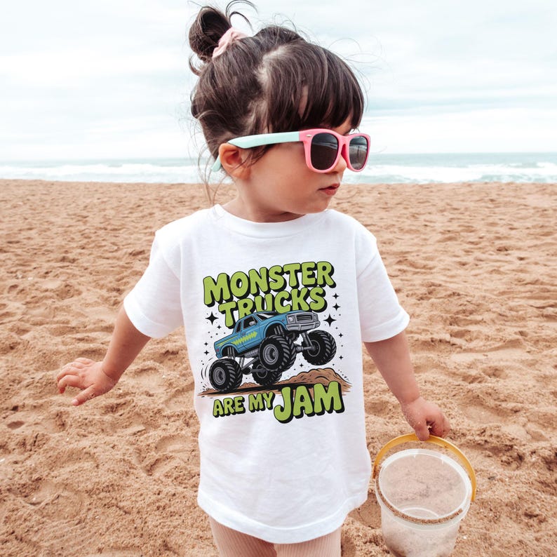 Puede incluir: Camiseta blanca con un gr&aacute;fico de un cami&oacute;n monstruo azul y el texto "MONSTER TRUCKS ARE MY JAM" en verde y negro. El ni&ntilde;o en la playa de arena lleva la camiseta y gafas de sol rosas.
