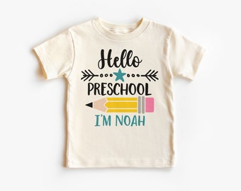 Puede incluir: Camiseta de manga corta color crema con el texto "Hello Preschool I'm Noah" en negro, turquesa y amarillo. El diseño incluye un lápiz y flechas, ideal para el primer día de escuela de un niño.