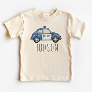 May include: A cream colored t-shirt with a blue and white police car illustration and the name "HUDSON" printed in black.