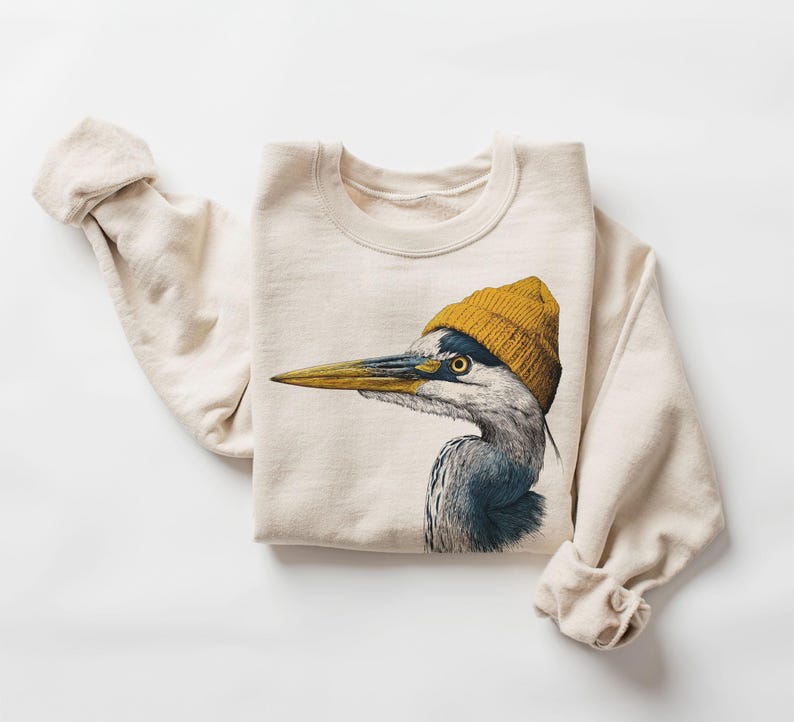 May include: A cream-coloured crewneck sweatshirt featuring a detailed graphic illustration of a grey heron wearing a mustard yellow knit beanie. The bird is shown in profile, facing left, with its long, pointed beak and textured plumage visible.