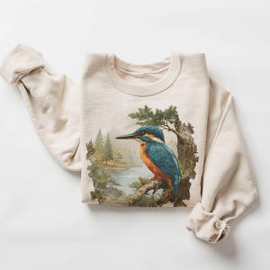 May include: Beige sweatshirt featuring a detailed illustration of a kingfisher bird perched on a branch. The bird has blue, orange, and white plumage. The background shows a landscape with trees and a body of water.