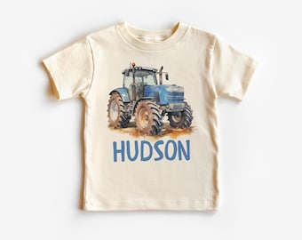 May include: Cream-colored short-sleeve t-shirt featuring a blue tractor illustration with brown tires. The name "HUDSON" is printed in blue block letters below the tractor. The shirt is made of soft fabric.