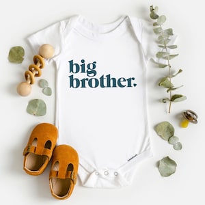 May include: White baby onesie with the text "big brother" in dark blue letters. The onesie is surrounded by eucalyptus leaves, a wooden rattle, and brown baby shoes.