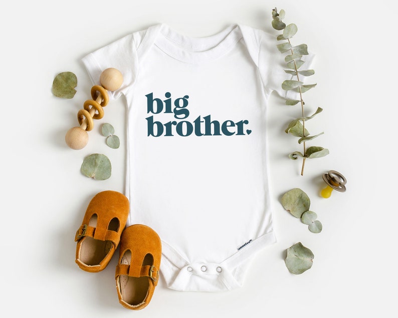 May include: White baby onesie with the text "big brother" in dark blue letters. The onesie is surrounded by eucalyptus leaves, a wooden rattle, and brown baby shoes.