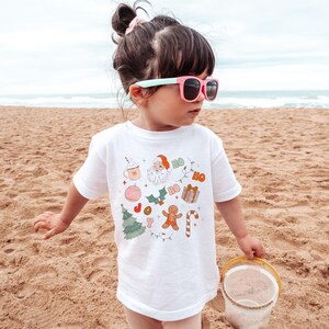 Puede incluir: Una camiseta blanca con un dise&ntilde;o navide&ntilde;o que presenta a Pap&aacute; Noel, un hombre de jengibre, adornos y la palabra "HO". La camiseta la lleva un ni&ntilde;o peque&ntilde;o en una playa de arena, con gafas de sol y un cubo.