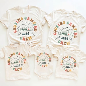 Könnte beinhalten: Fünf cremefarbene T-Shirts und ein Body mit einem Camping-Motiv. Das Design enthält die Worte "Cousins Camping Crew 2026" in einem kreisförmigen Muster um eine Berglandschaft mit einem Wohnwagen, Bäumen und Marshmallows.