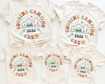 Può includere: Cinque magliette e un body color crema con una grafica a tema campeggio. Il design include le parole "Cousins Camping Crew 2026" in un motivo circolare attorno a una scena di montagna con un camper, alberi e marshmallow.