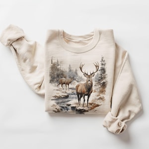May include: A beige sweatshirt with a graphic print of two deer in a snowy forest setting. The deer are brown with antlers and are standing in a stream. The background is a watercolor-style painting of trees and snow.