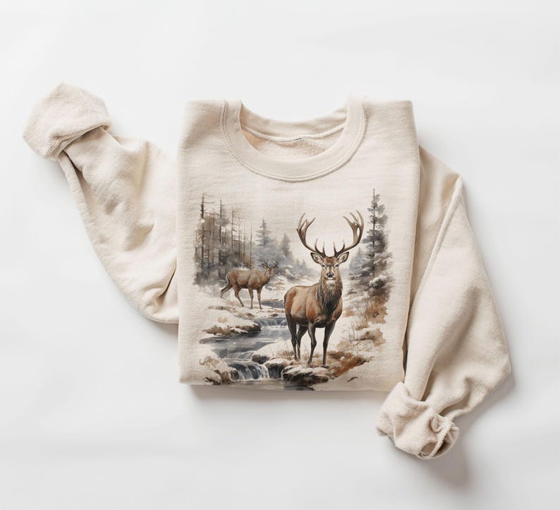 May include: A beige sweatshirt with a graphic print of two deer in a snowy forest setting. The deer are brown with antlers and are standing in a stream. The background is a watercolor-style painting of trees and snow.