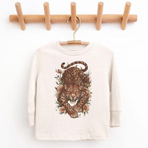 Puede incluir: Una camiseta de manga larga color crema con una ilustración detallada de un leopardo rodeado de flores. El leopardo tiene un patrón de manchas en tonos marrones y beige. La camiseta está colgada en una percha de madera.