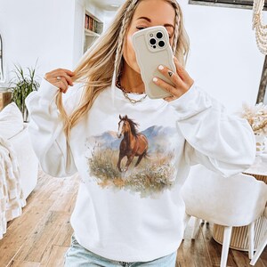 May include: White sweatshirt featuring a watercolor painting of a brown horse running through a field with mountains in the background. The horse is the focal point, with detailed brushstrokes. The sweatshirt has long sleeves and a crew neck.
