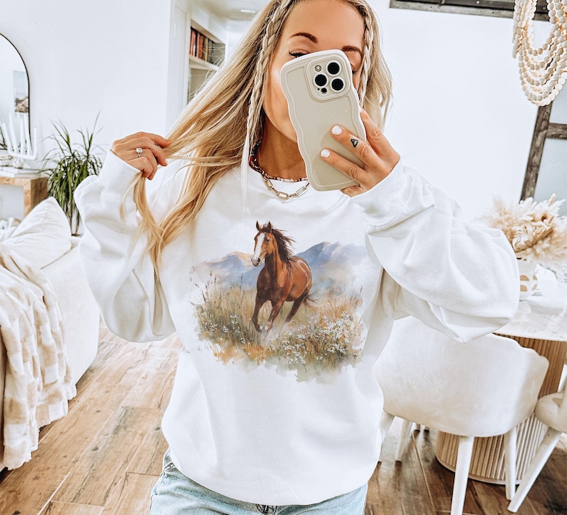 May include: White sweatshirt featuring a watercolor painting of a brown horse running through a field with mountains in the background. The horse is the focal point, with detailed brushstrokes. The sweatshirt has long sleeves and a crew neck.